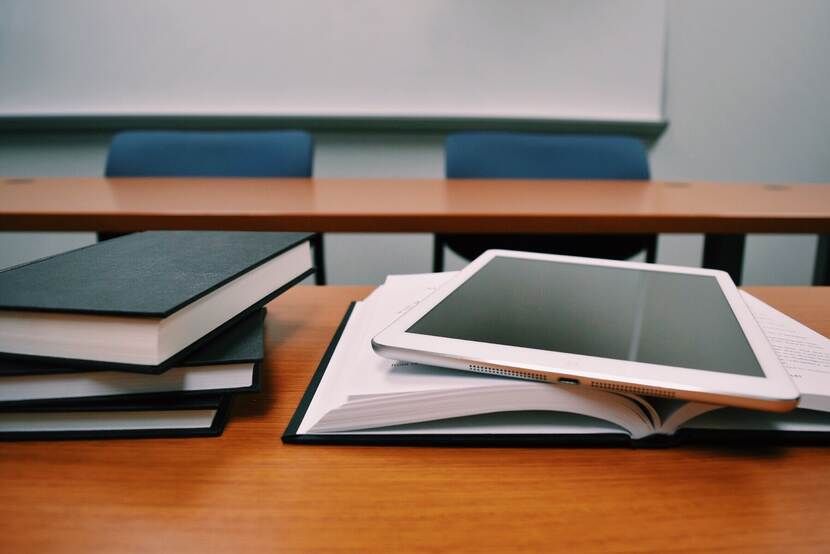 Twee bureau's met stoelen erachter. Op het voorste bureau ligt links een stapel boeken en rechts een open boek met hierop een tablet.