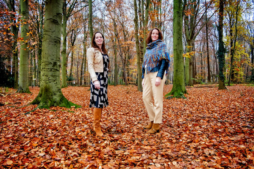 Twee vrouwen staan in een bos. Onder hen is de grond bedekt met oranje bladeren